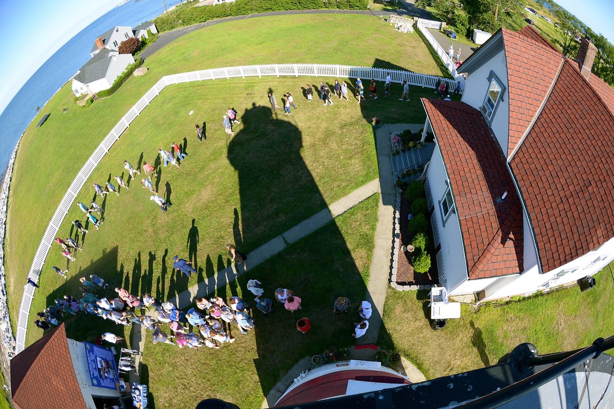 Fisheye view from the top of Stratford Point Lighthouse, its shadow across the lawn, visitors in a circle below and the water off to the left