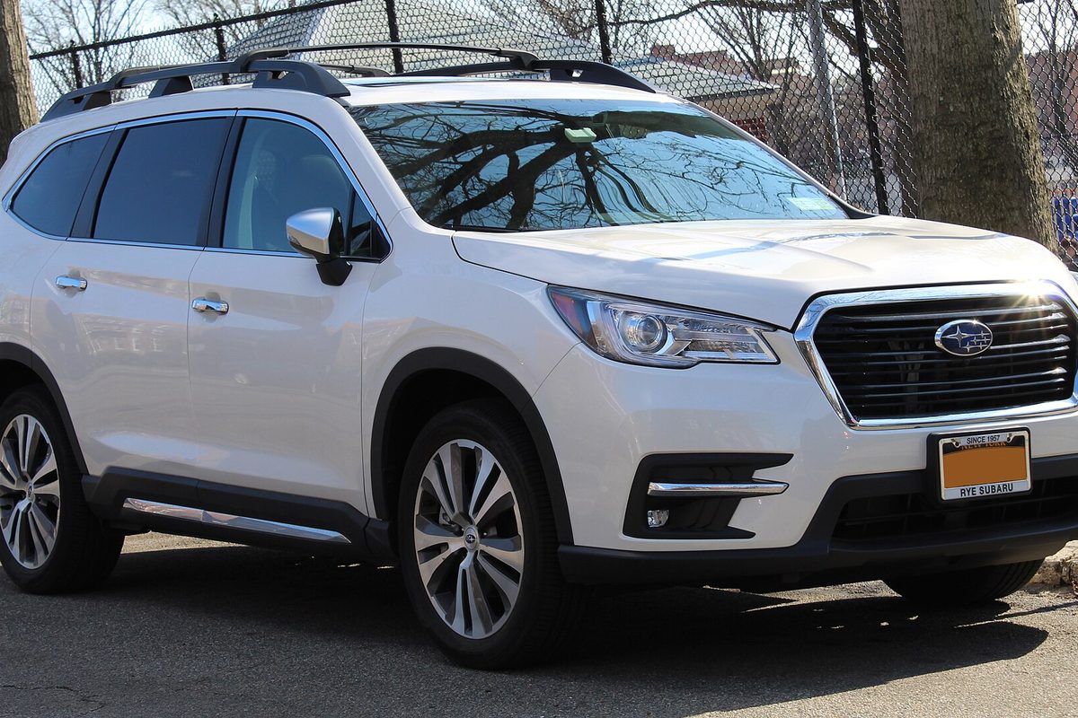 A white Subaru Ascent SUV with roof rails, parked by a chain-link fence and bare trees