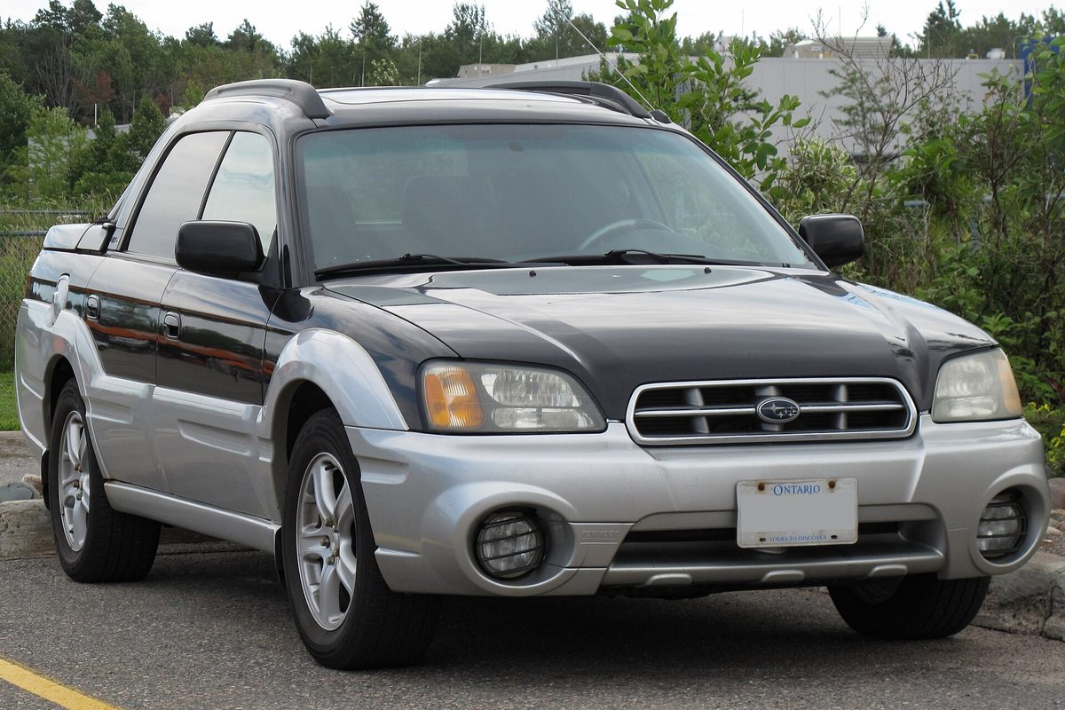 A two-tone silver and black 2003 Subaru Baja with roof rails, front three-quarter view, parked on a lot with trees behind
