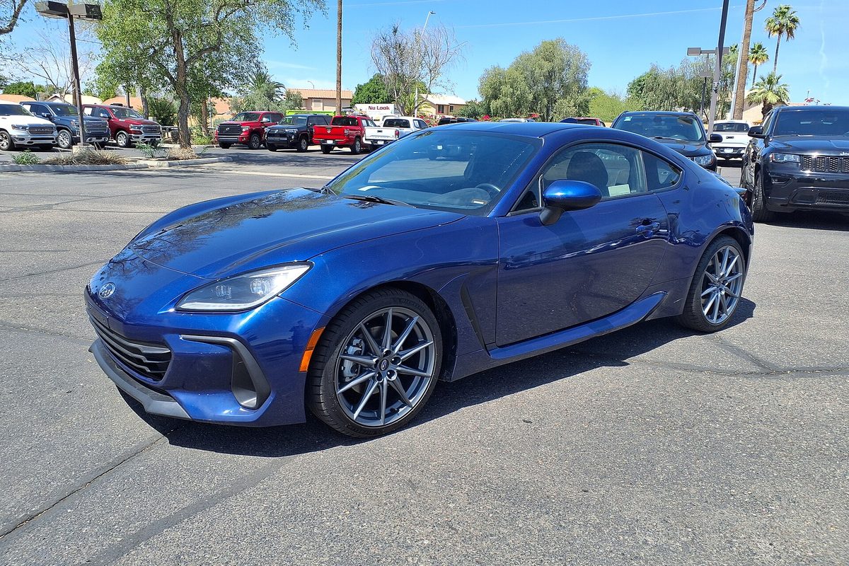 A blue 2023 Subaru BRZ coupe with silver alloy wheels on a sunny parking lot, palm trees and pickups behind it