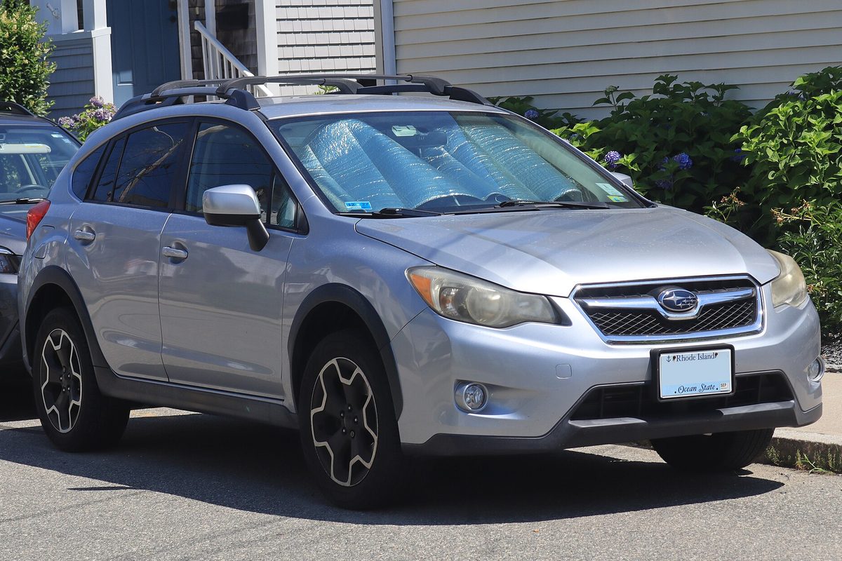A silver 2014 Subaru XV Crosstrek with black wheels and a sunshade in the windshield, parked by a house