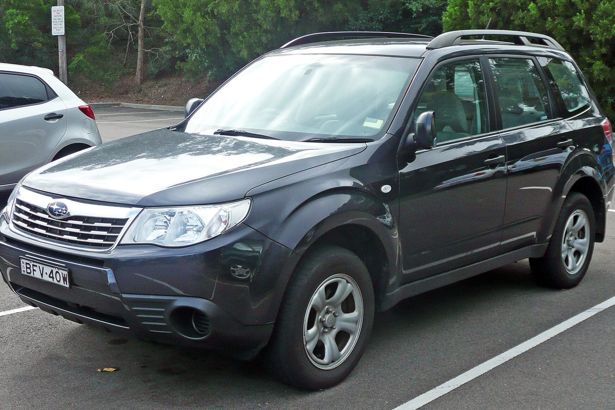 A dark gray 2008 Subaru Forester parked in a lot with trees and a white car behind it