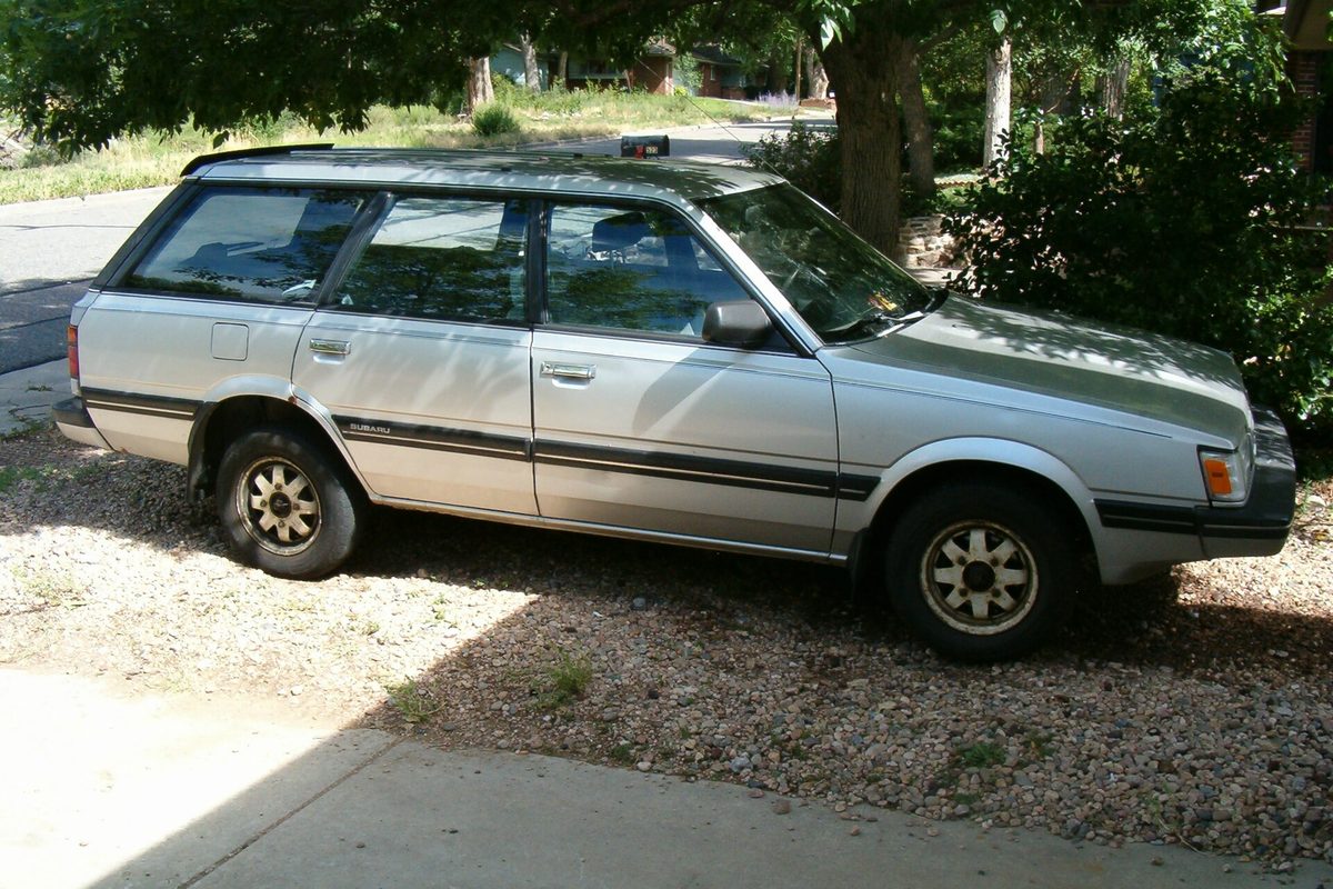 A silver 1988 Subaru GL wagon with a dark side stripe and gold-tone wheels, parked on a gravel driveway in the shade of a tree