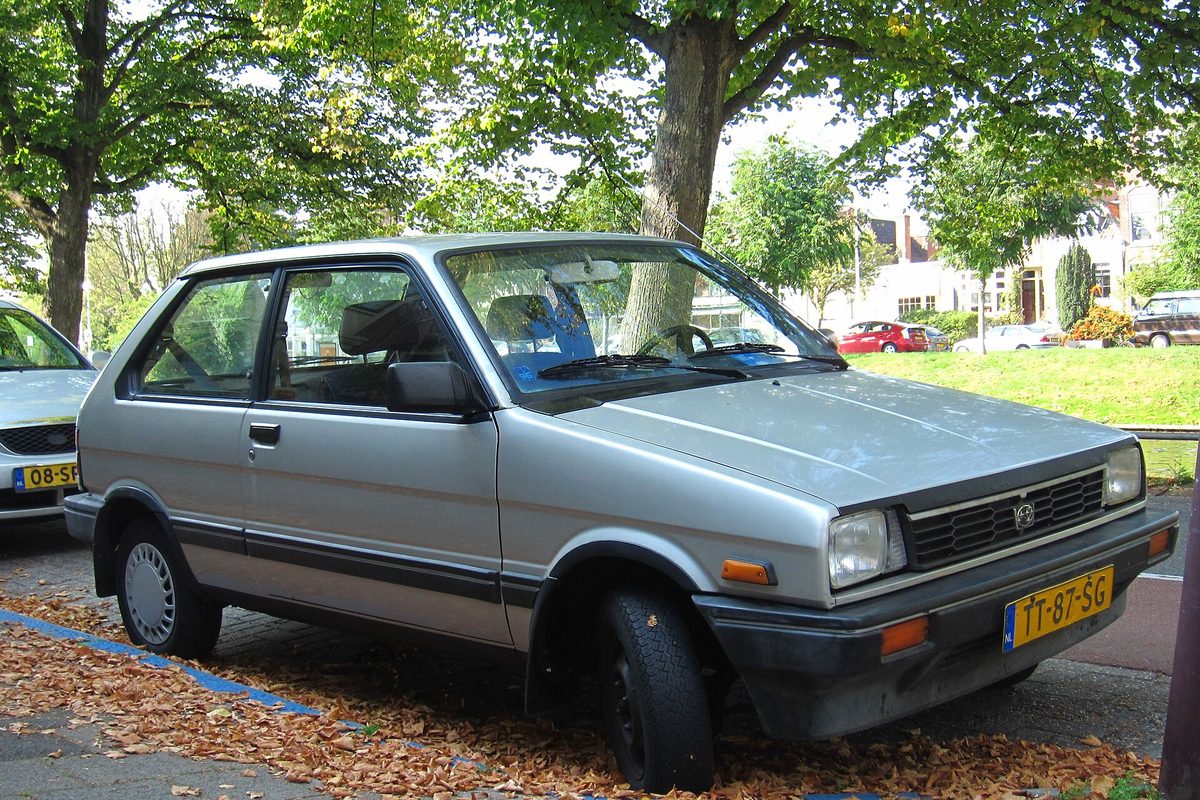 A silver three-door Subaru Justy hatchback parked on fallen leaves under trees on a city street