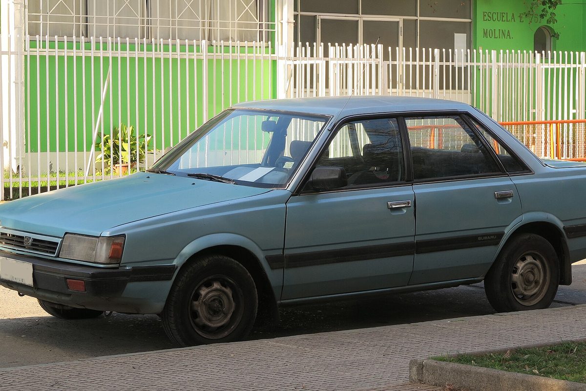 A faded light blue Subaru Loyale four-door sedan parked at a curb in front of a white fence and a green building