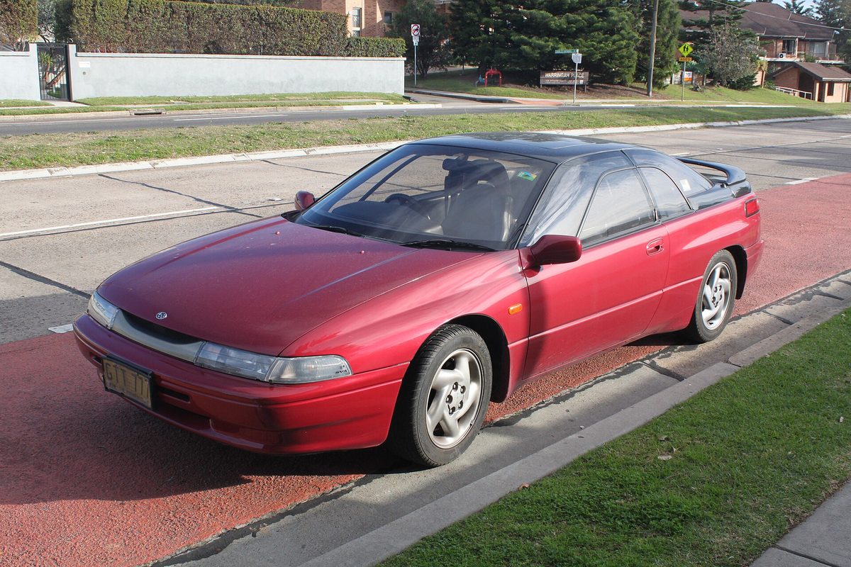 A dark red Subaru SVX coupe with its black glass roofline and silver alloy wheels, parked at a curb beside a grass verge