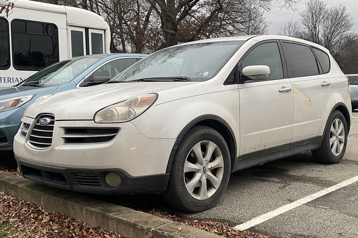 A dirty white 2006 Subaru B9 Tribeca SUV with clouded headlights and a scuffed rear door, parked in a lot with fallen leaves