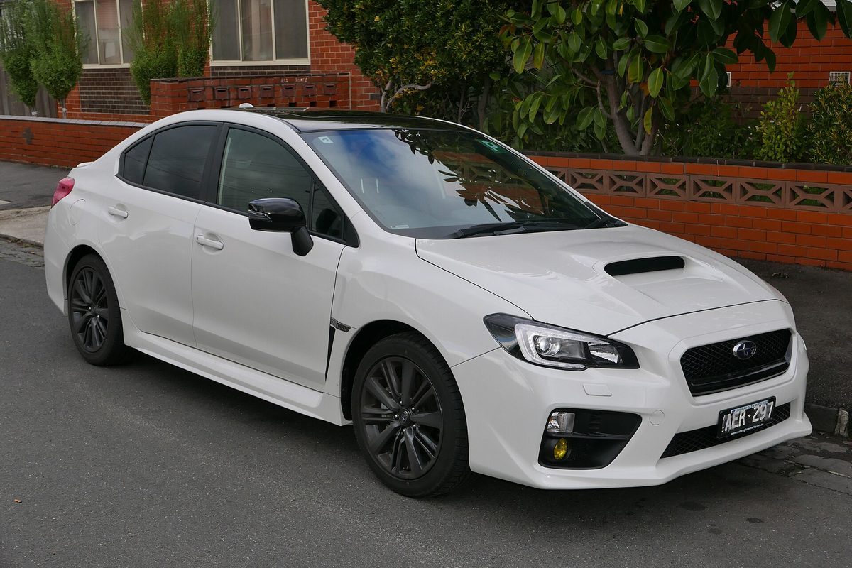 A white Subaru WRX sedan with black wheels and a hood scoop, parked at a curb in front of a red brick wall
