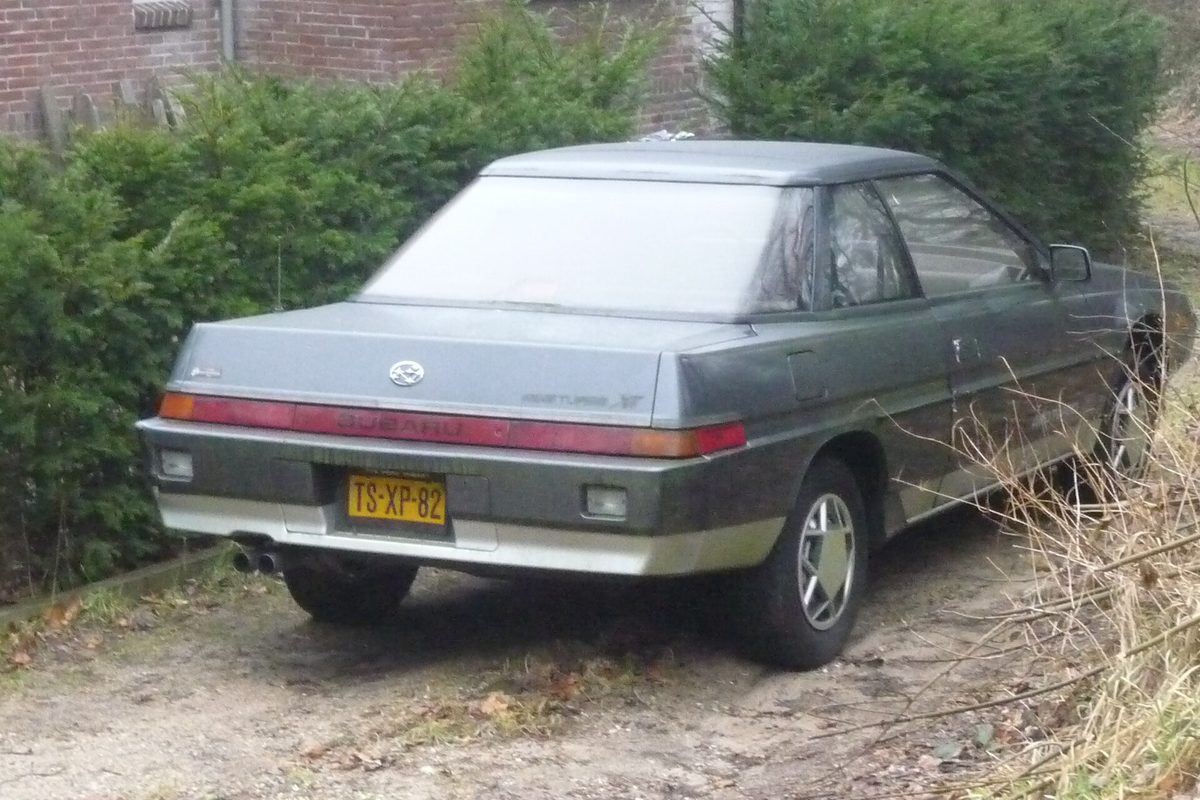 Rear view of a gray two-tone Subaru XT coupe parked on a gravel drive beside a hedge and a brick wall