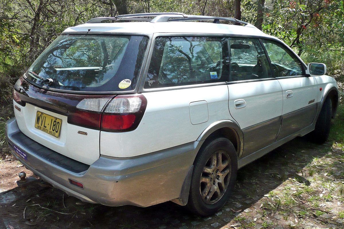 A white 2000 Subaru Outback wagon with gray lower body cladding, seen from the rear three-quarter angle, parked on dirt among trees in Audley, New South Wales, Australia