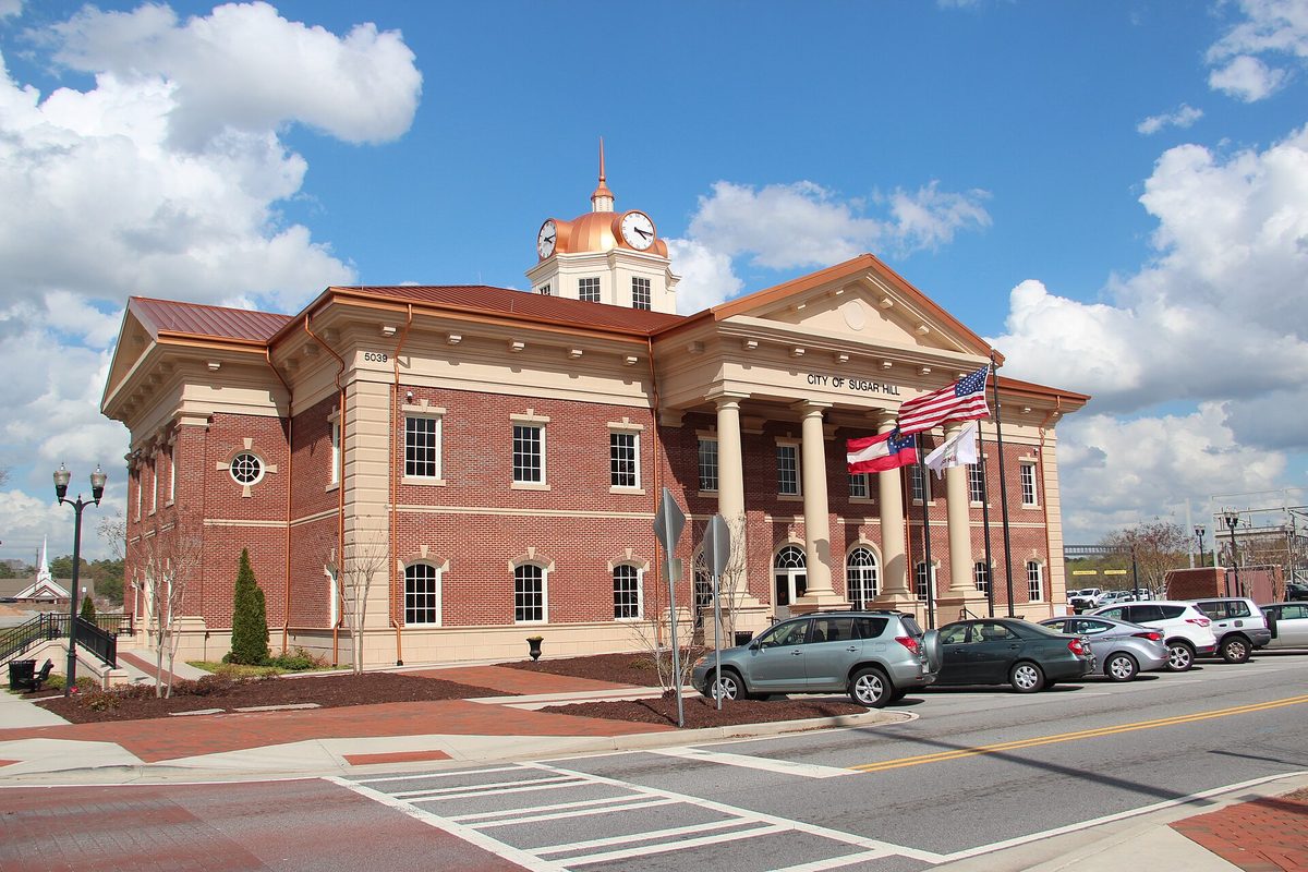 Sugar Hill City Hall, a red-brick building with white columns and a copper-domed clock cupola, with cars parked along the street in front