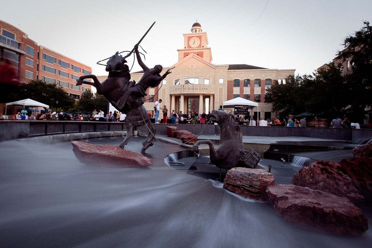 A bronze statue of a rider on a rearing horse in the fountain at Sugar Land Town Square, with the clock-towered City Hall and a crowd under white tents behind