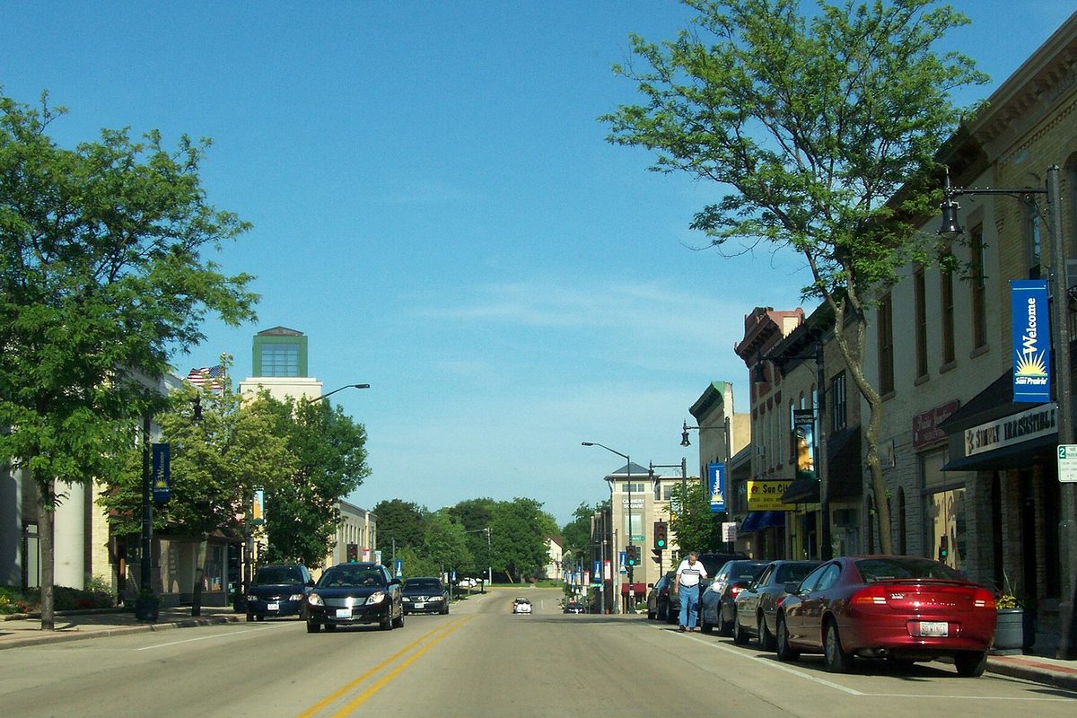 Main Street in downtown Sun Prairie on a sunny day, with brick storefronts, cars parked at the curb and a Welcome to Sun Prairie banner on a light pole