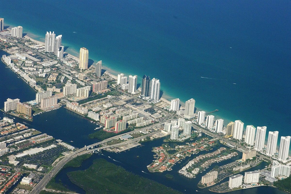 Aerial view of a narrow barrier island lined with high-rise towers between the blue ocean and a bay of canals, with a bridge crossing to the mainland