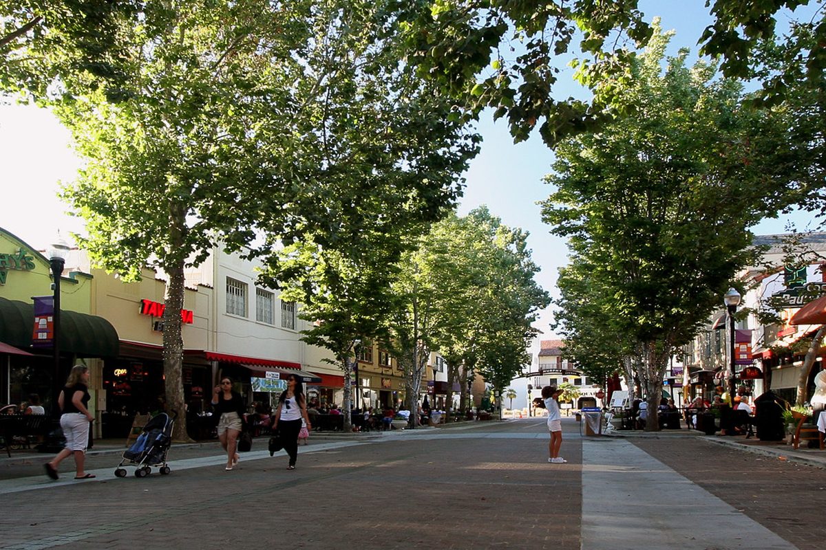 People walking down a brick pedestrian street in Sunnyvale lined with leafy trees and restaurant awnings
