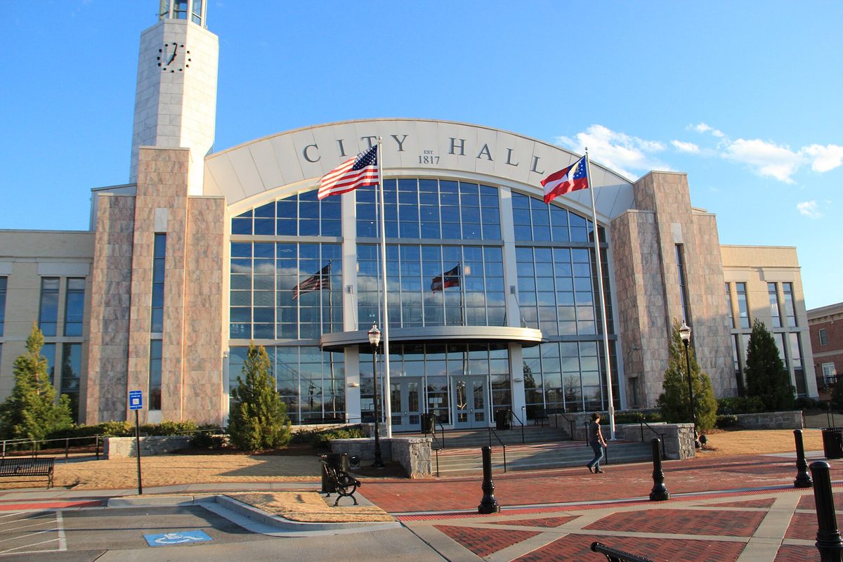 Suwanee City Hall with a curved glass front, a stone clock tower and United States and Georgia flags, under a late-afternoon blue sky