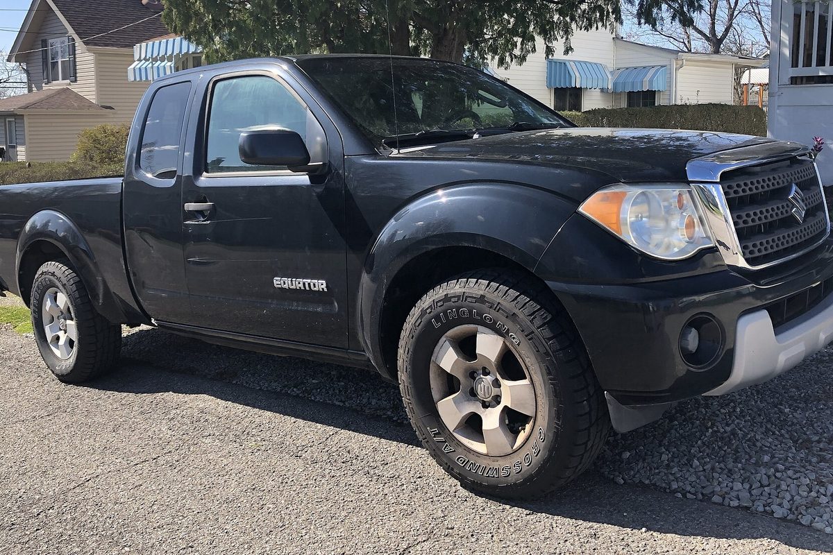 A black Suzuki Equator extended-cab pickup on all-terrain tires, parked on a gravel driveway in front of houses