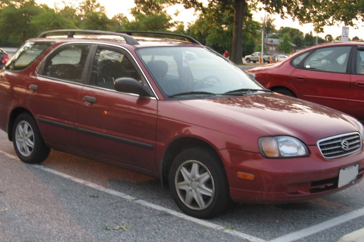 A dark red Suzuki Esteem wagon with roof rails and silver wheels, parked in a parking lot under trees
