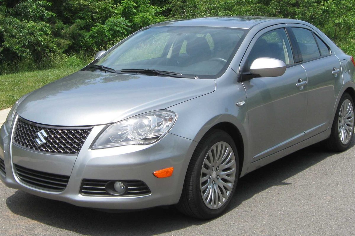 A silver Suzuki Kizashi sedan with multi-spoke alloy wheels, parked on a road with green trees behind it