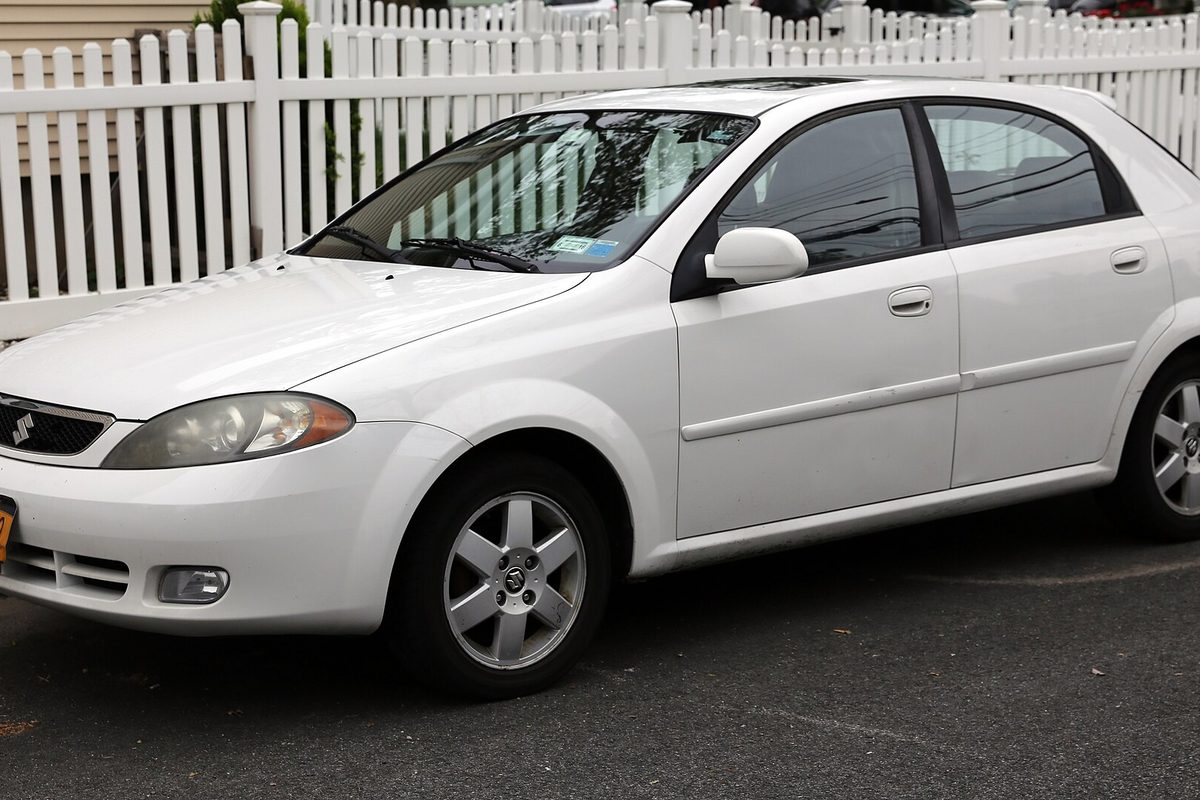 A white Suzuki Reno five-door hatchback with silver alloy wheels, parked in front of a white picket fence