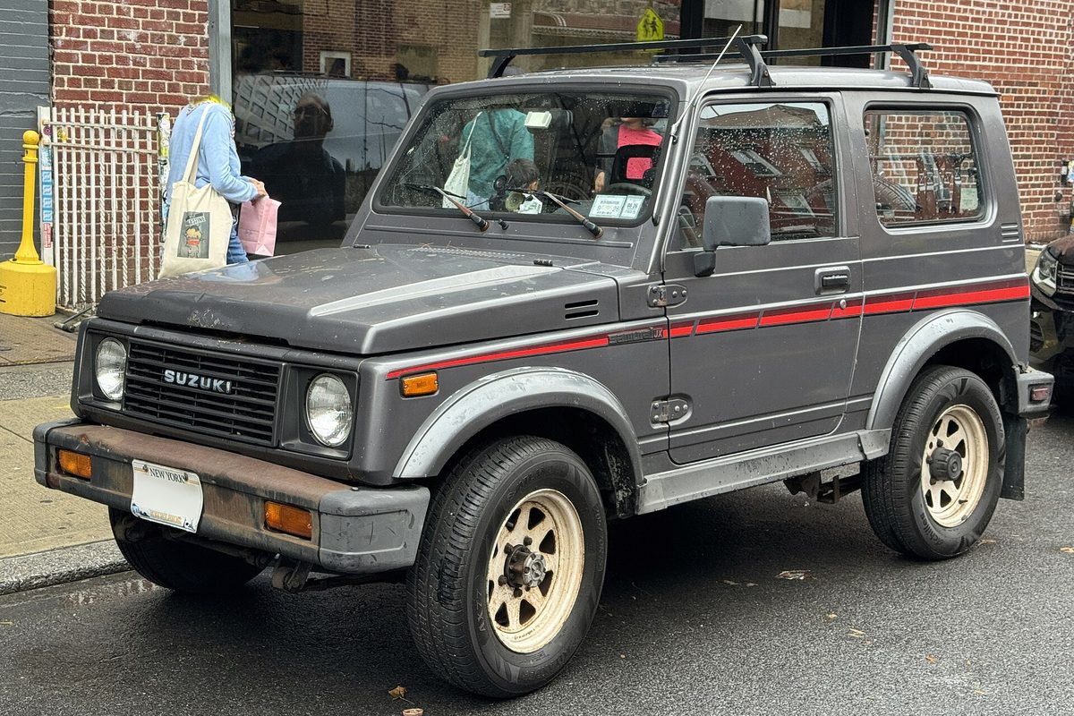 A gray two-door Suzuki Samurai hardtop with a red side stripe, roof rack and tan steel wheels, parked on a city street