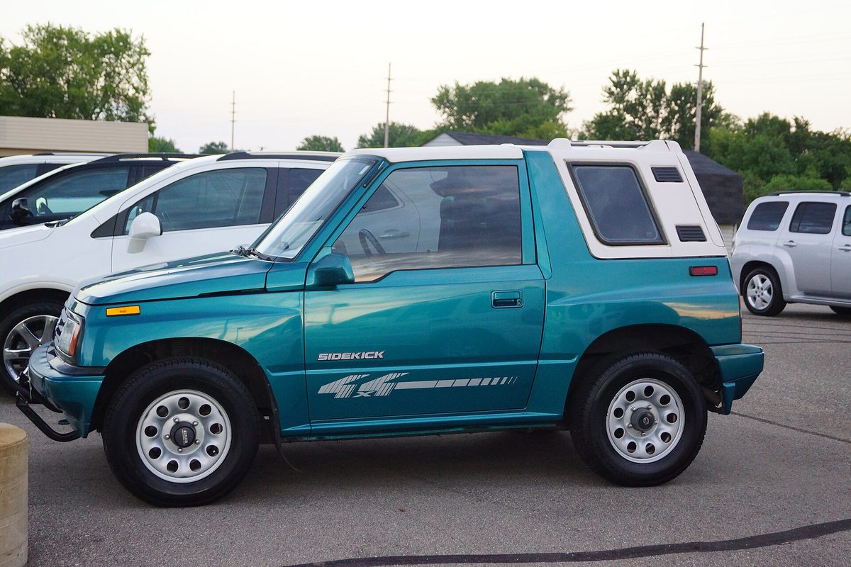 A teal two-door Suzuki Sidekick with a white hardtop and Sidekick side decals, seen from the side in a parking lot