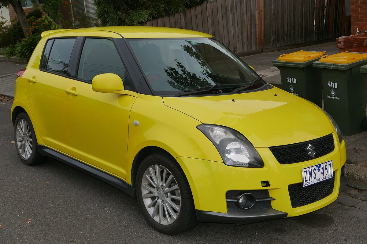 A bright yellow Suzuki Swift Sport five-door hatchback with silver alloy wheels, parked at a curb beside two wheelie bins