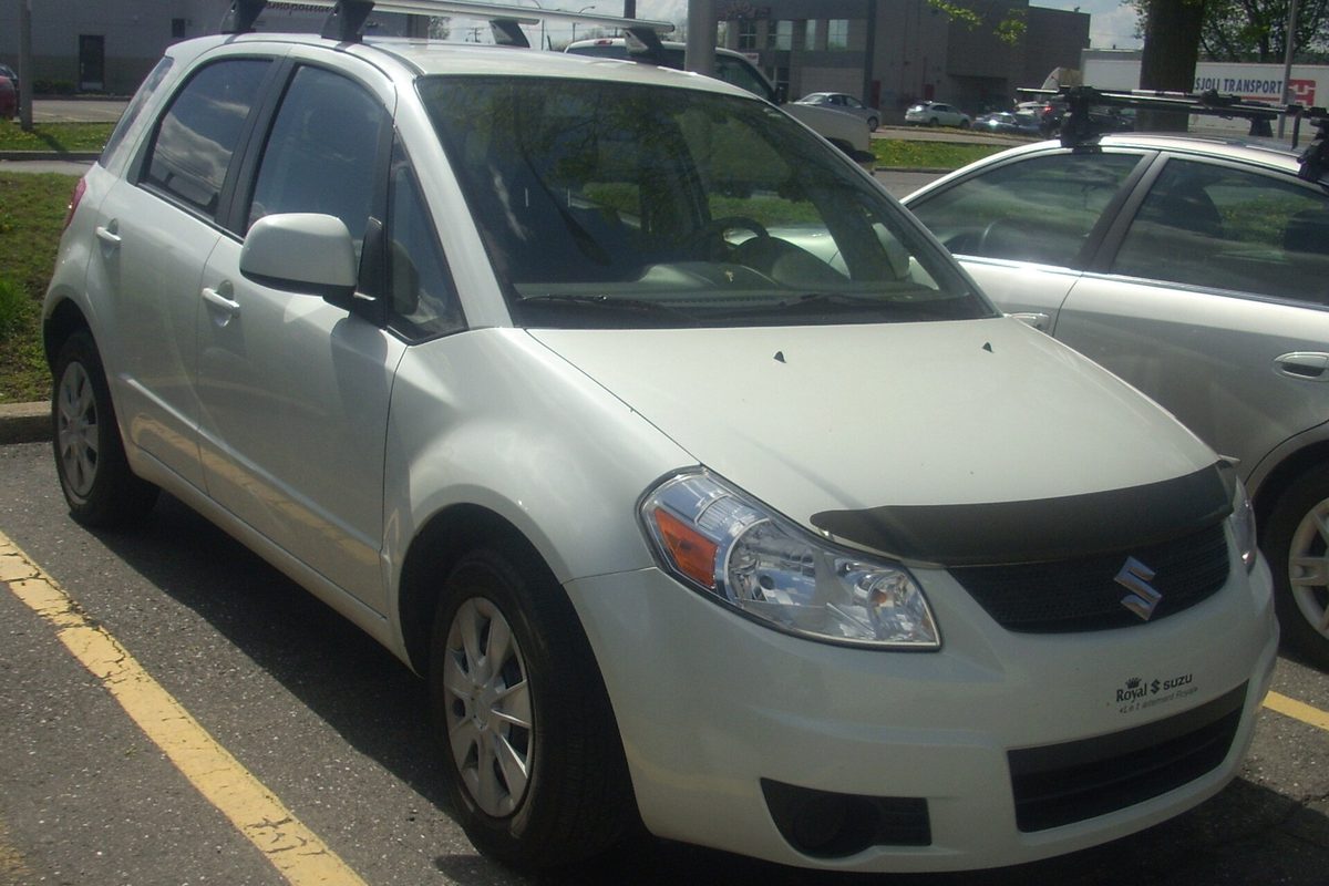 A pearl-white Suzuki SX4 hatchback with roof rack bars and a hood deflector, parked in a lot