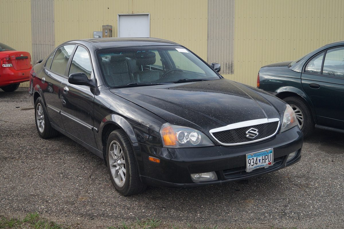 A dusty black Suzuki Verona sedan parked on gravel beside a tan metal building