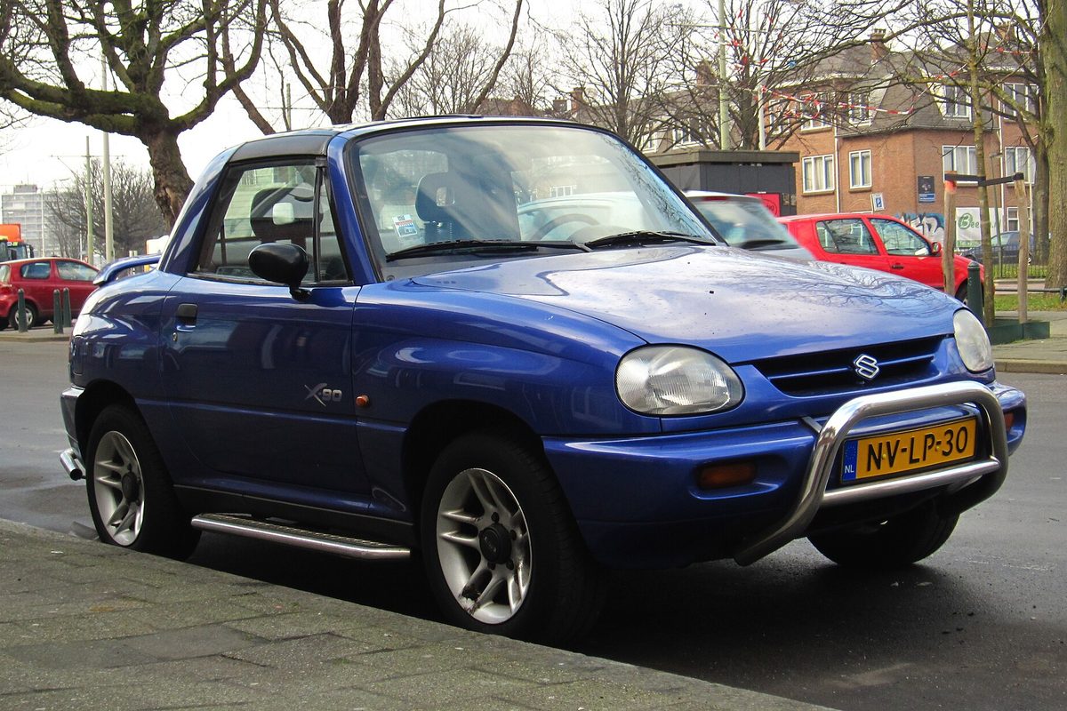 A blue Suzuki X-90 two-door with a black roof, chrome front bar and side steps, parked on a city street with bare trees behind it