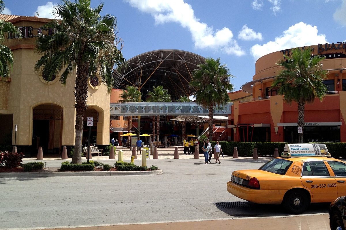 The arched entrance of Dolphin Mall under a blue sky, with palm trees, shoppers on the plaza and a yellow taxi at the curb