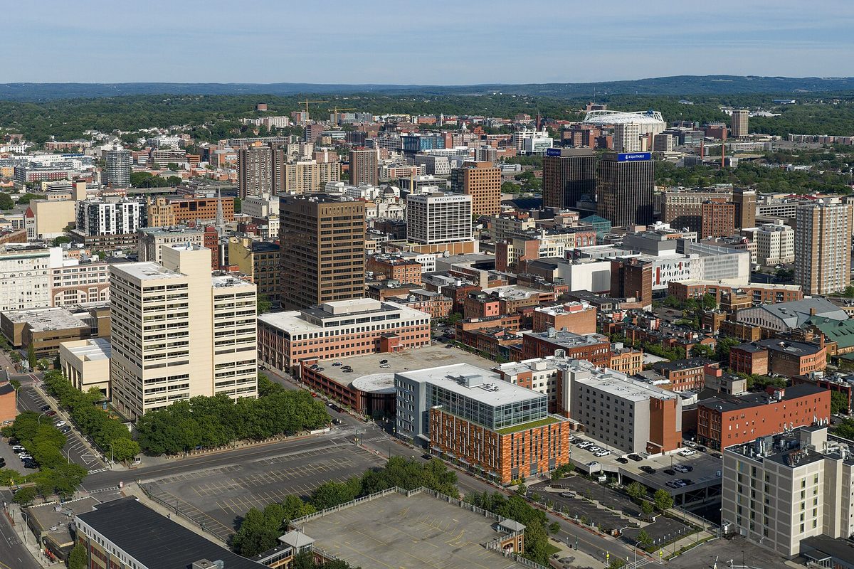 Aerial view of downtown Syracuse, with office towers and brick buildings among green trees and low hills on the horizon