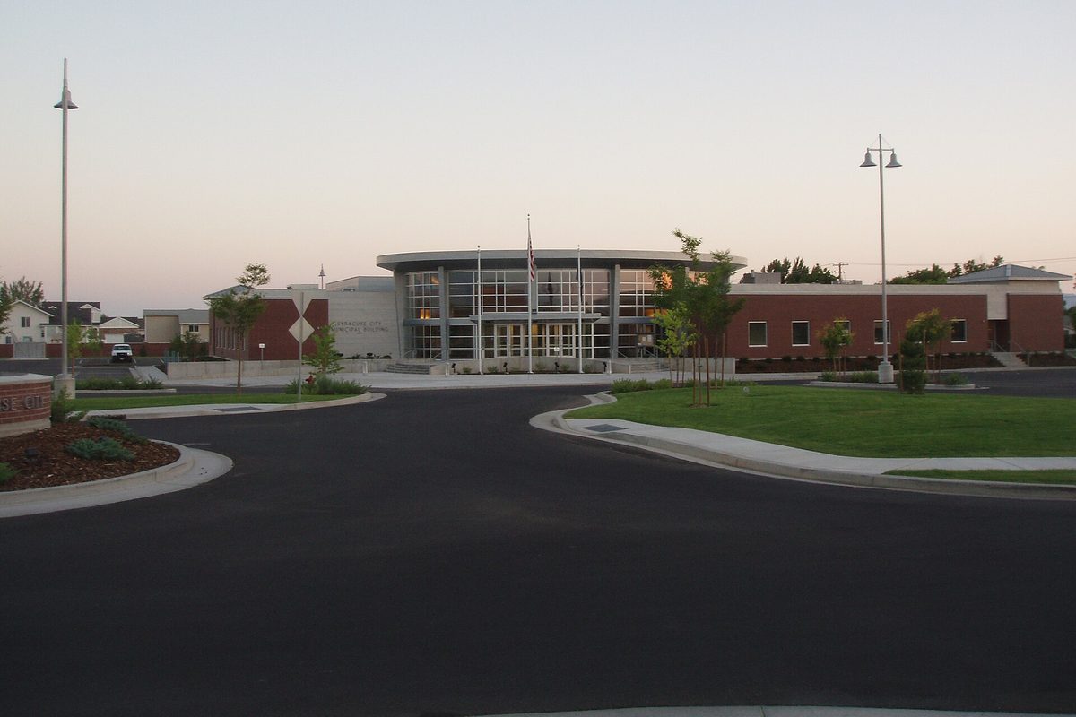 The Syracuse City Municipal Center at dusk, a brick building with a curved glass entrance, seen across a curving driveway and lawn