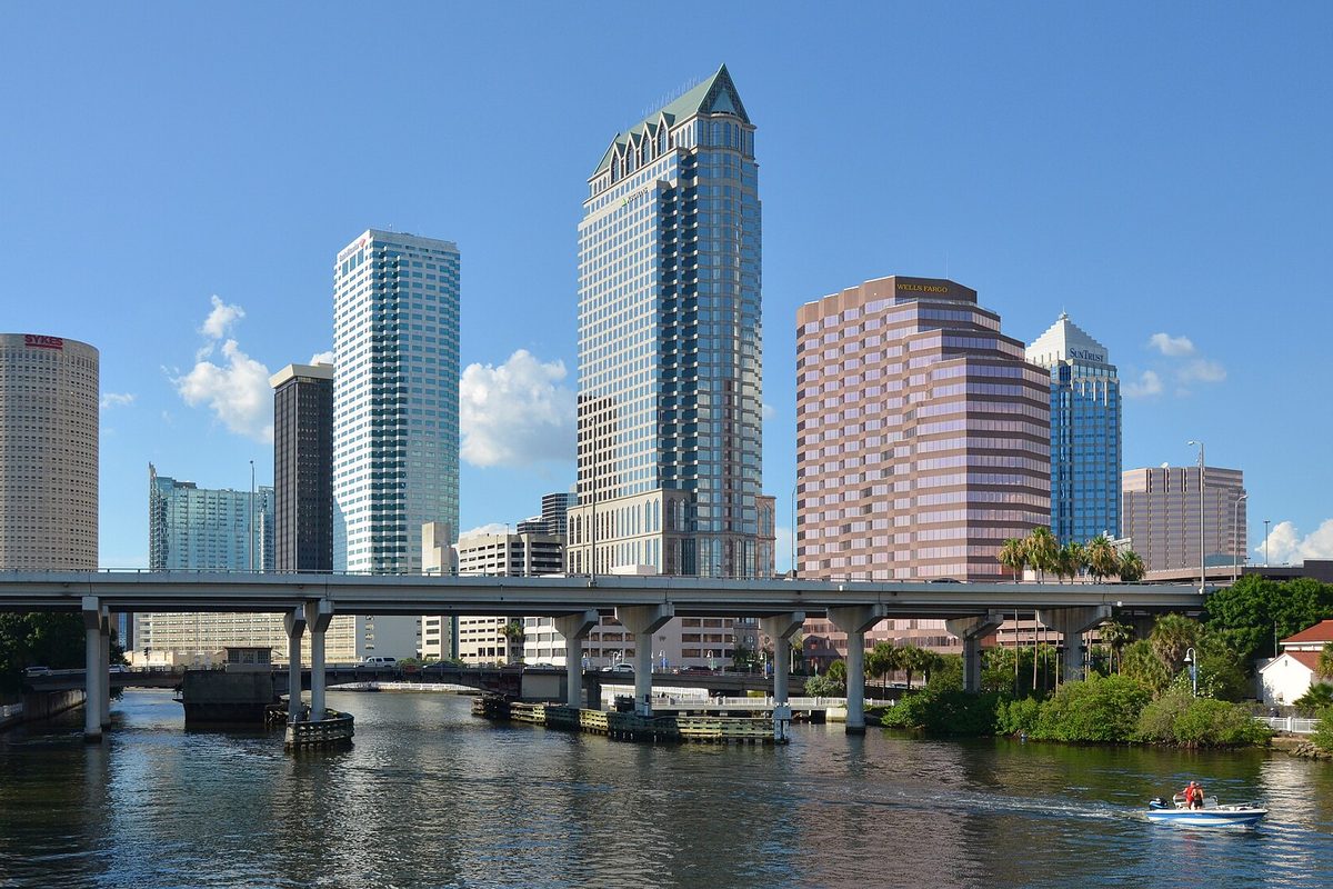 Downtown Tampa glass towers seen across the Hillsborough River, with a highway bridge crossing the water and a small motorboat in front