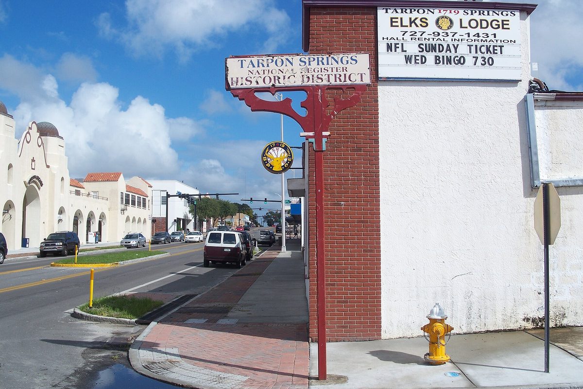 A red signpost reading Tarpon Springs National Register Historic District on a downtown corner, beside the Elks Lodge wall, with Spanish-style buildings and parked cars down the street