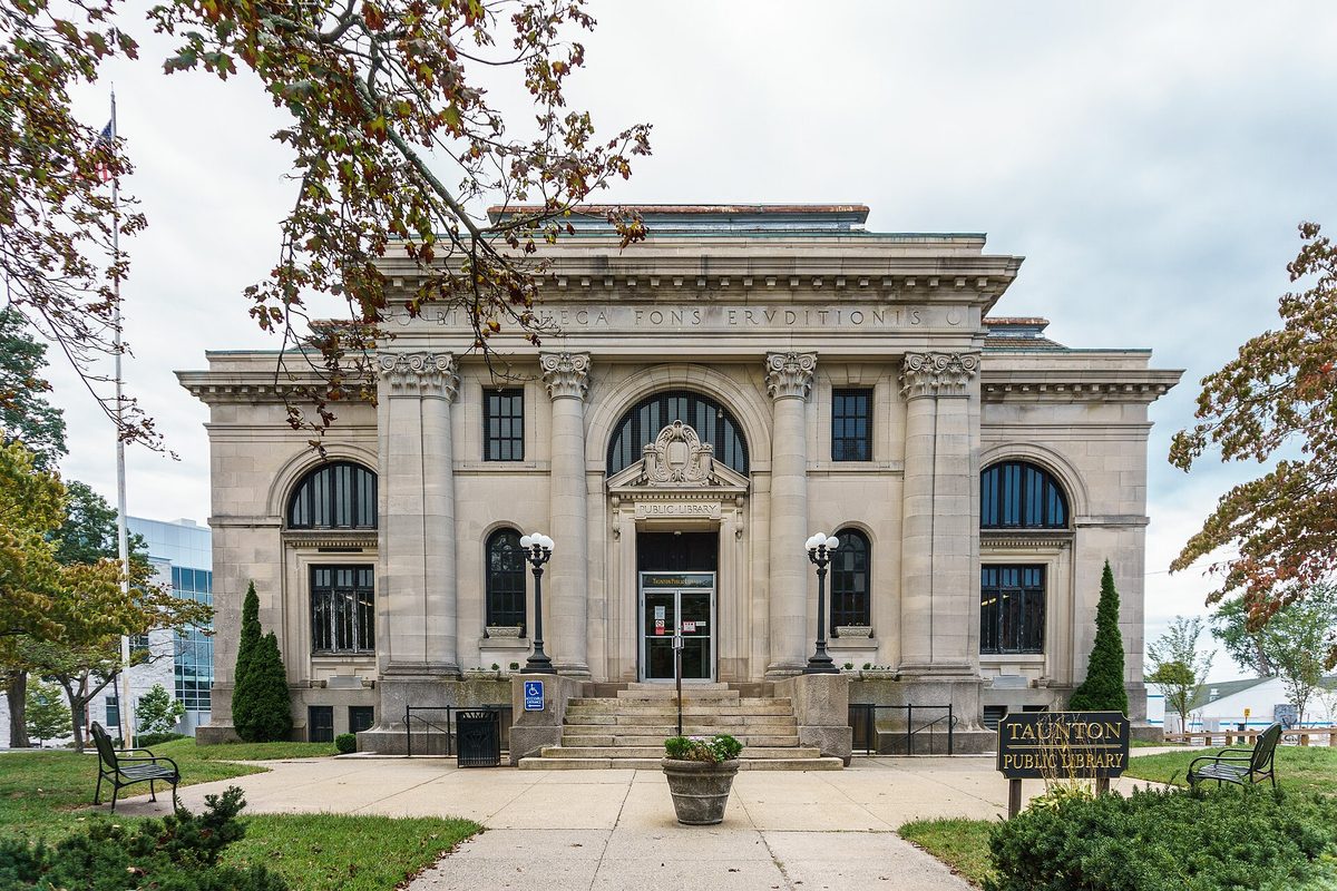 The columned stone front of the Taunton Public Library, with its name sign on the lawn and benches beside the front walk
