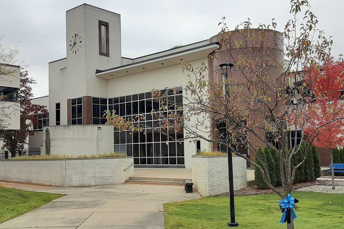 Taylor Municipal Offices, a white building with a clock on its tower and a wall of tall windows, behind a lawn with autumn trees and a blue ribbon on a lamp post