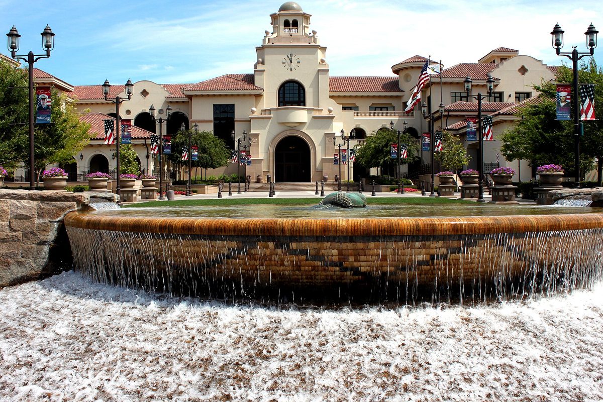 Temecula City Hall, a cream building with a clock tower and red tile roofs, lined with American flags and lamp posts, behind a round cascading fountain