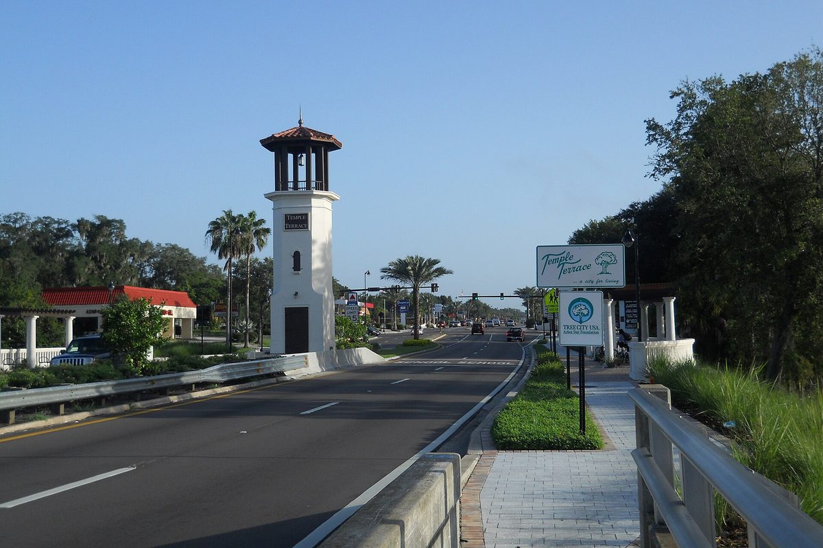 A white entry tower with a red tile cupola beside 56th Street in Temple Terrace, with palm trees, a city welcome sign and the road running into the distance