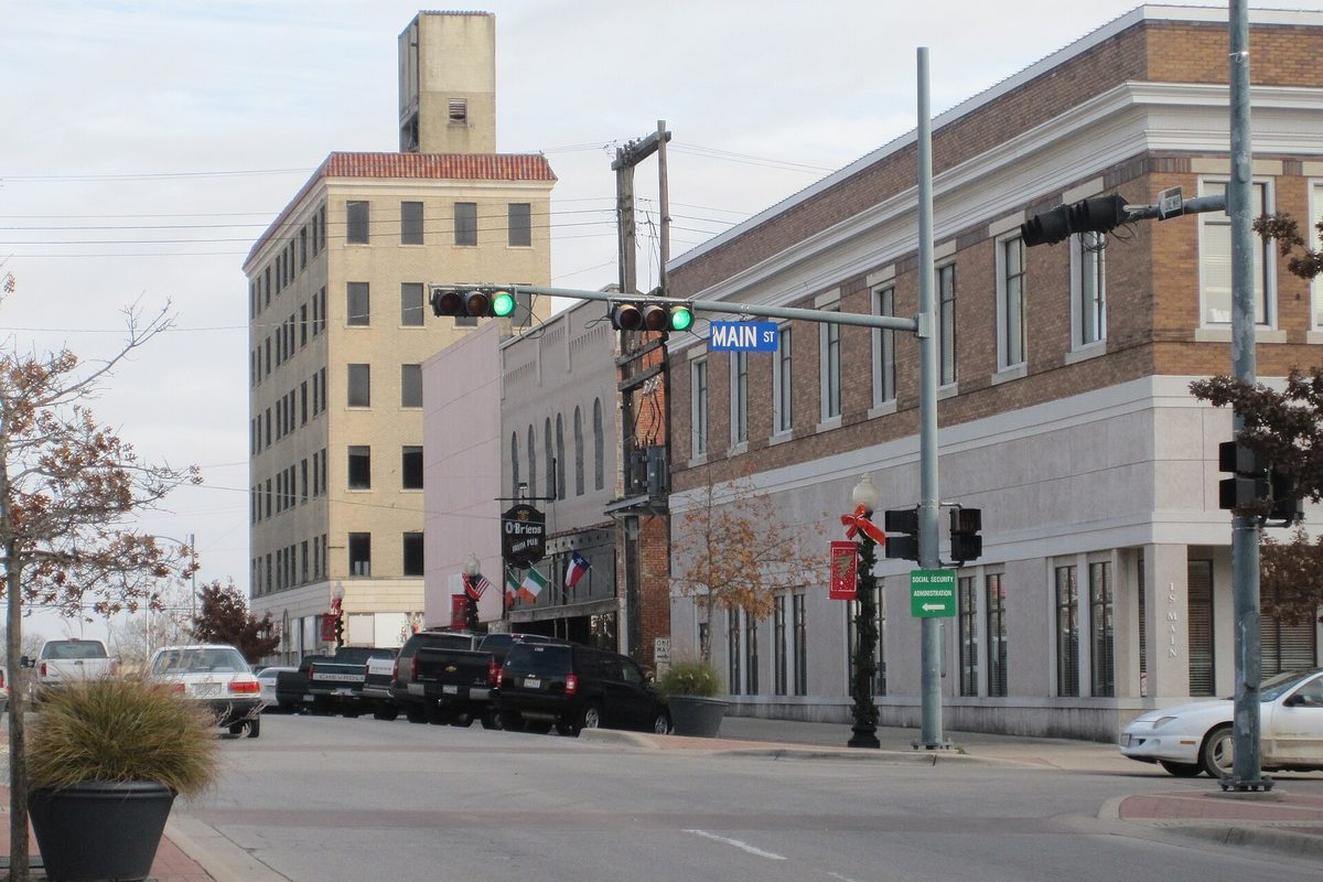 Main Street in downtown Temple at an intersection with traffic lights, a tall cream-colored building with a red tile roof and a brick corner building, pickup trucks parked along the curb