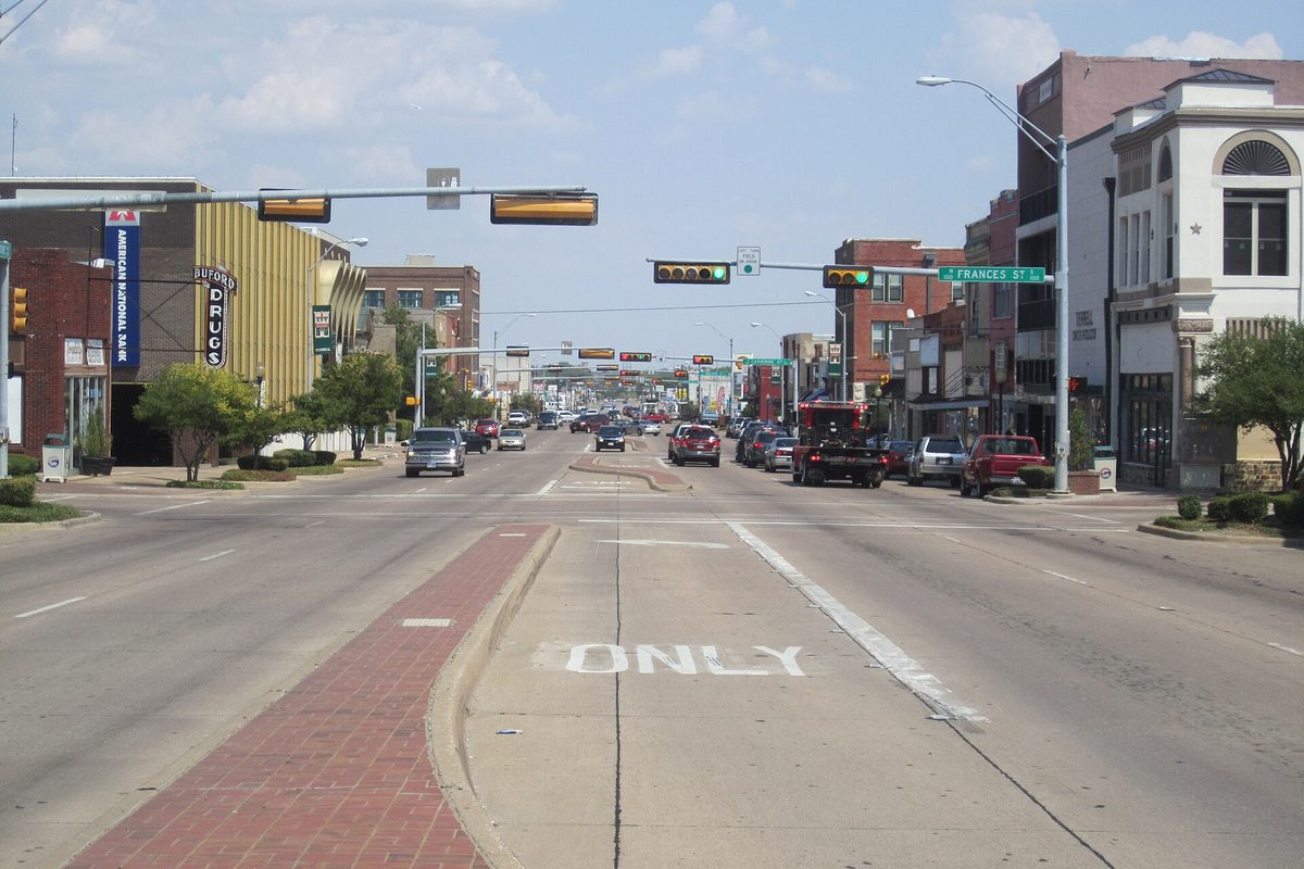 U.S. Highway 80 running through downtown Terrell, a wide street with traffic lights, brick storefronts, an old drugstore sign and parked pickups
