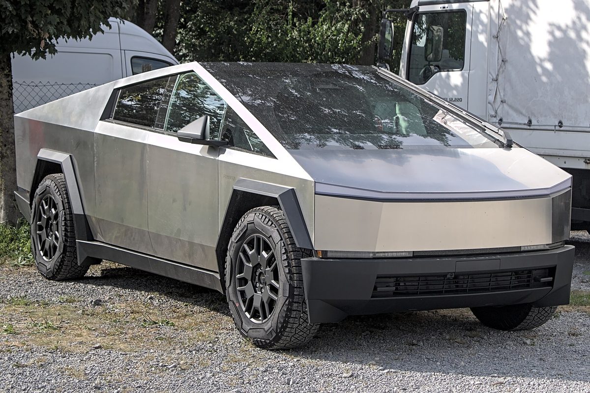 A brushed stainless-steel Tesla Cybertruck with black wheels and off-road tires, parked on gravel in front of a white truck