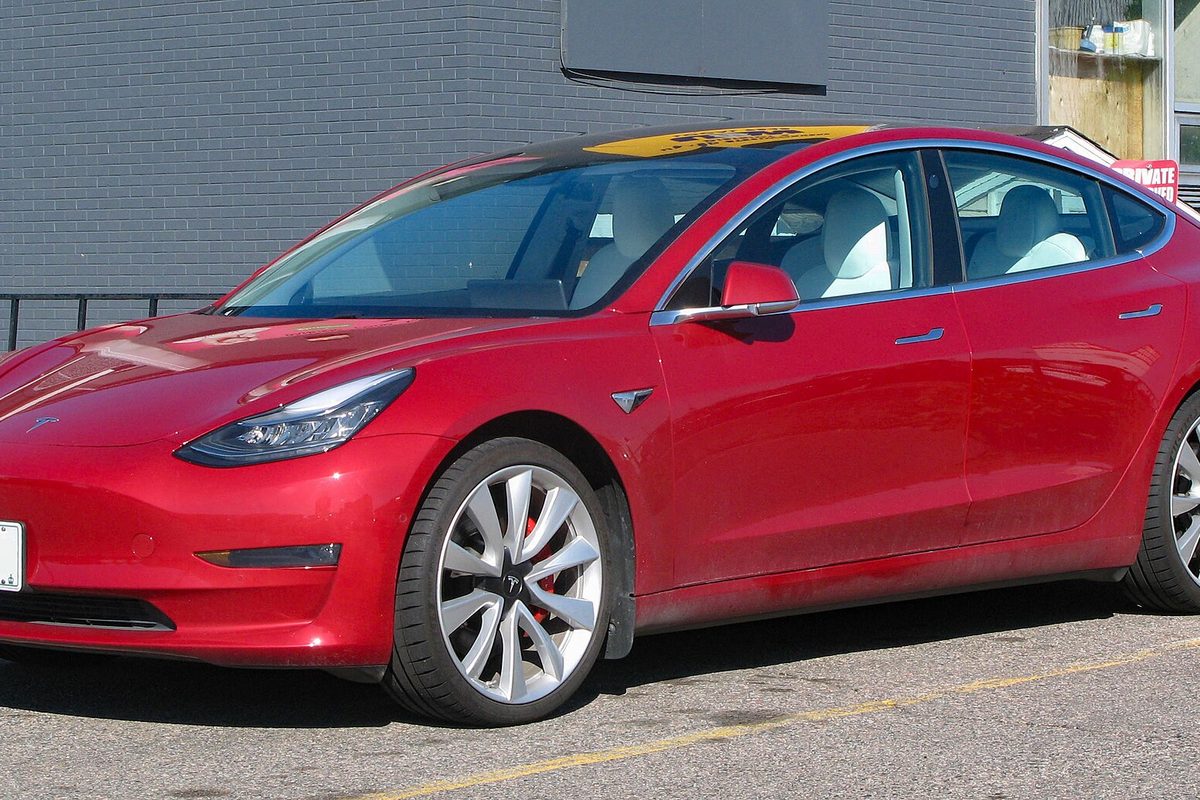 A red Tesla Model 3 sedan with a white interior and silver wheels, parked on asphalt in front of a gray brick wall