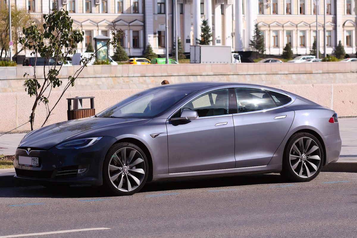 A dark gray Tesla Model S sedan parked at a curb in front of a stone wall and a large white columned building