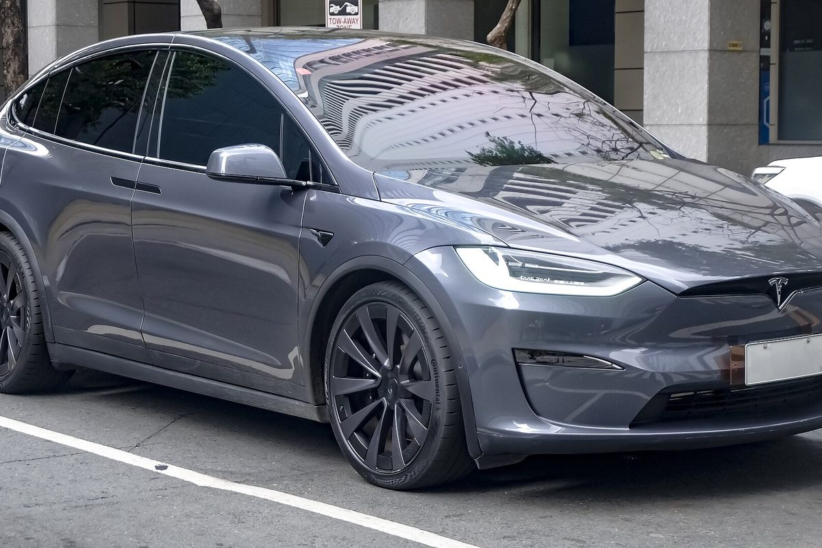 A dark gray Tesla Model X SUV with black wheels, parked at a painted curb beside tall stone pillars