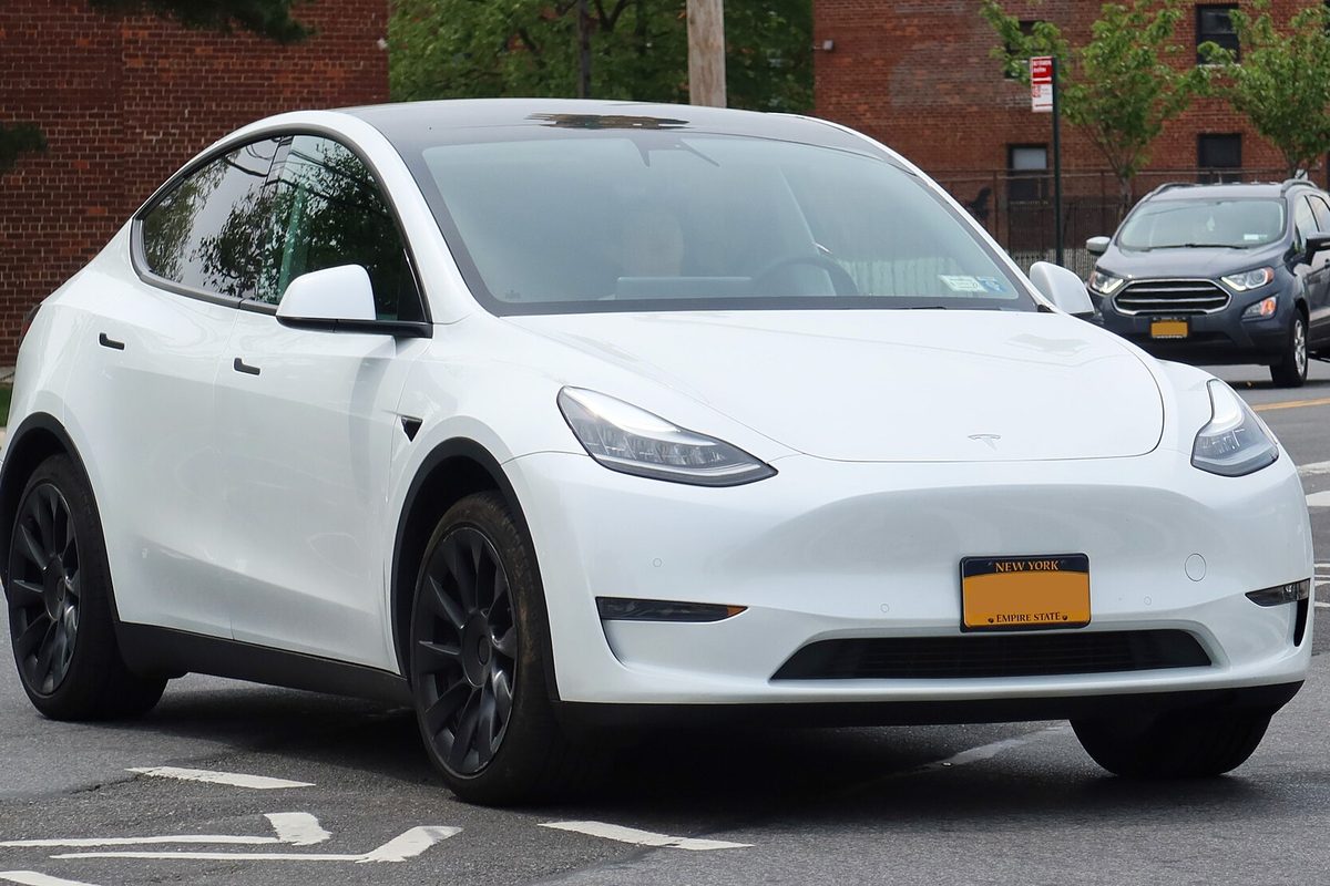 A white Tesla Model Y SUV with black wheels on a city street, brick buildings and a parked SUV behind it