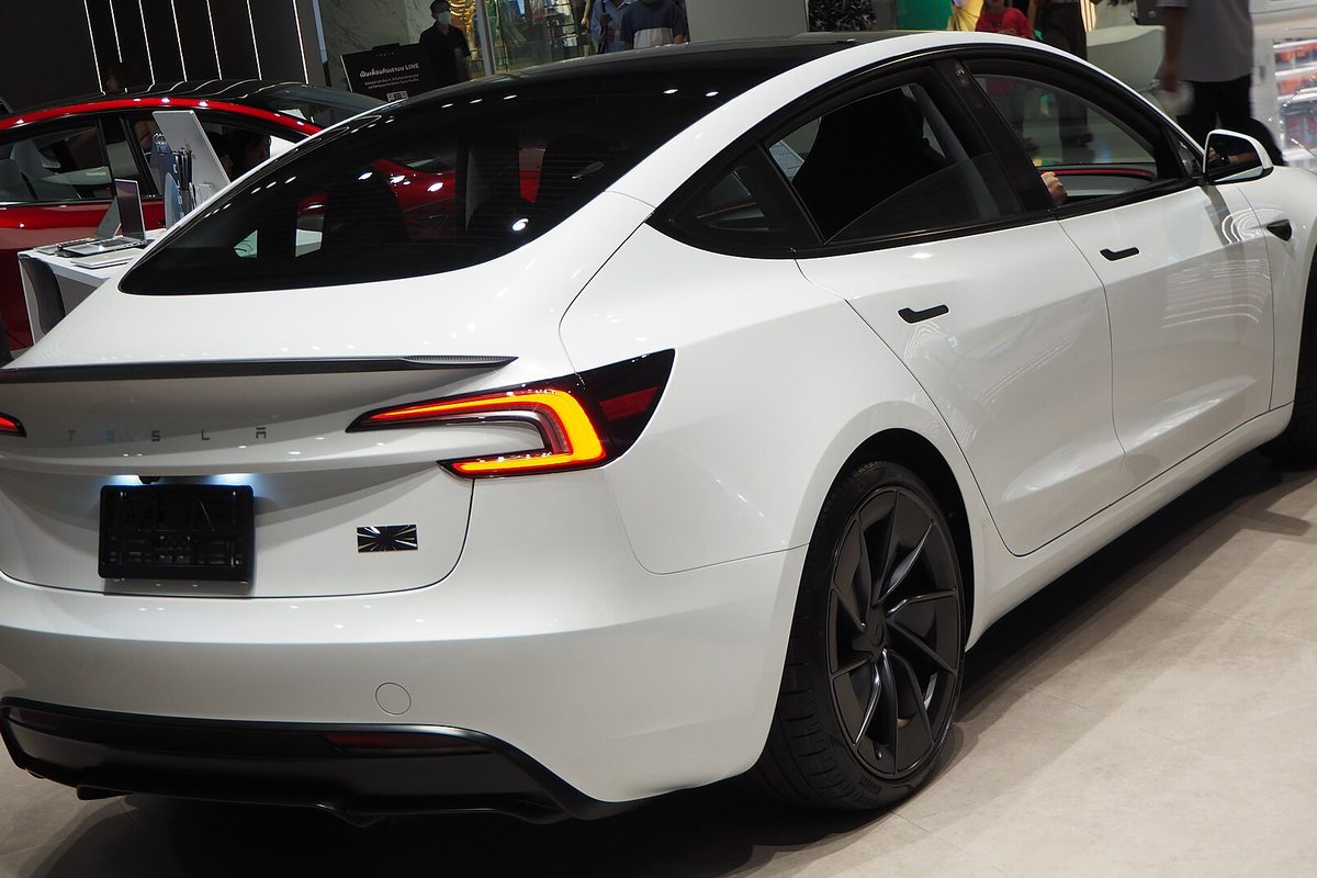 Rear three-quarter view of a white 2024 Tesla Model 3 Performance with a black rear spoiler and dark wheels, parked indoors at a display in Bangkok