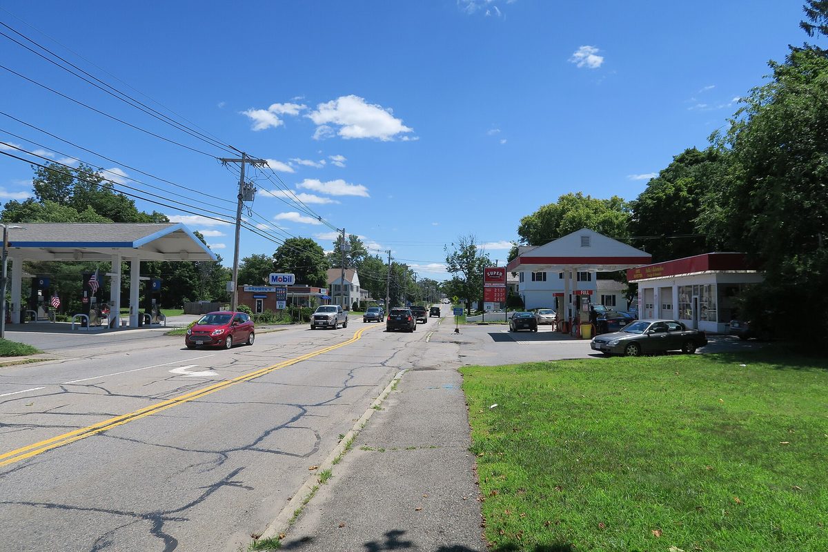 Main Street in Tewksbury on a summer day, a two-lane road with a gas station canopy on the left, a small red-and-white gas station on the right and cars driving away