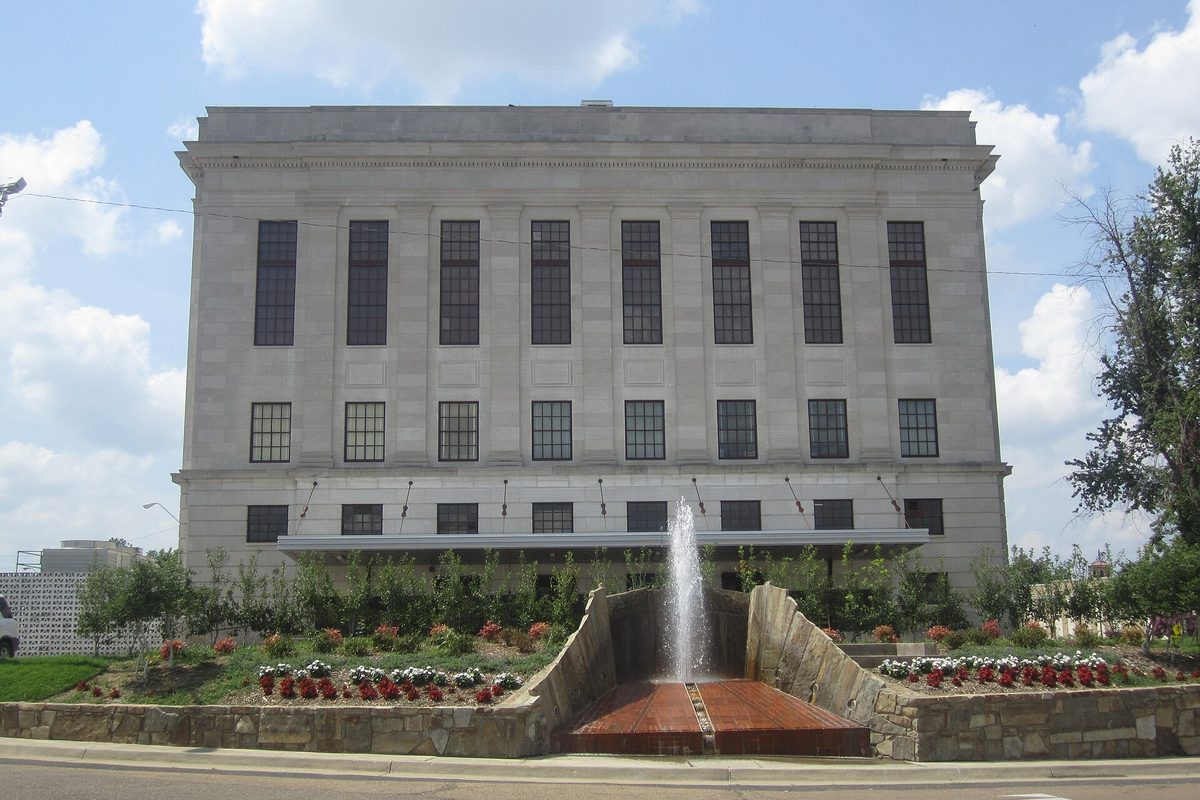 The pale stone back of the U.S. Courthouse in Texarkana, with a fountain and beds of red and white flowers in front, under a blue sky with white clouds