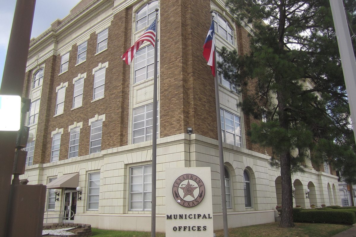 The brick-and-stone Texarkana municipal building with an American flag and a Texas flag out front, beside a sign with the city seal reading Municipal Offices